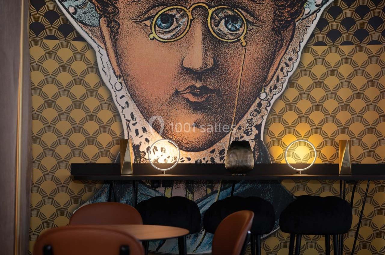 Illustration murale d'un visage stylisé avec des lunettes, décorée dans un intérieur avec des lampes et des chaises noires.