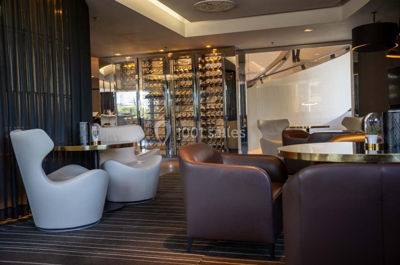 Salon moderne avec fauteuils en cuir, tables rondes et mur vitré exposant des bouteilles de vin.