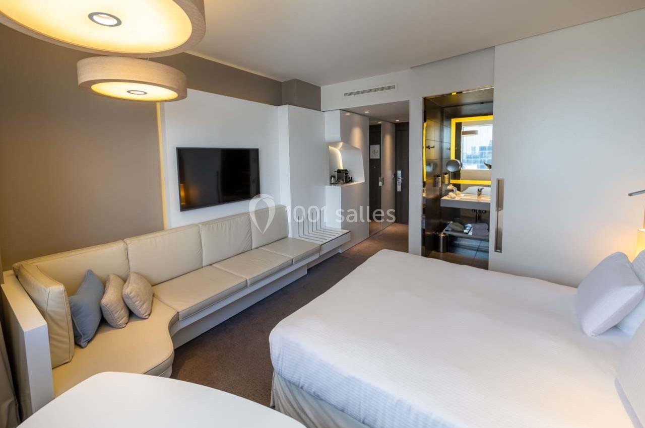 Chambre d'hôtel moderne avec lit double, canapé, télévision murale et salle de bain visible à travers une paroi vitrée.