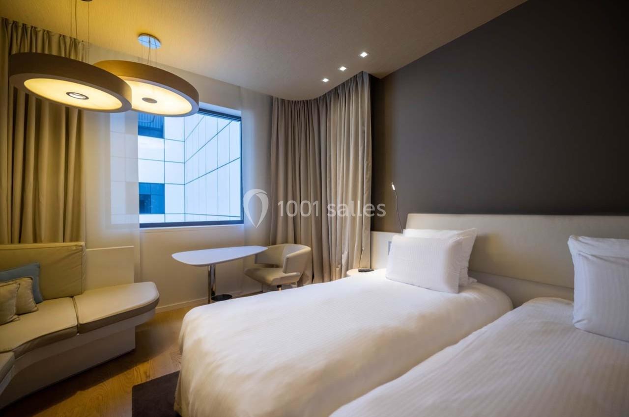 Chambre moderne avec deux lits simples, un coin salon, une table et une grande fenêtre laissant entrer la lumière naturelle.