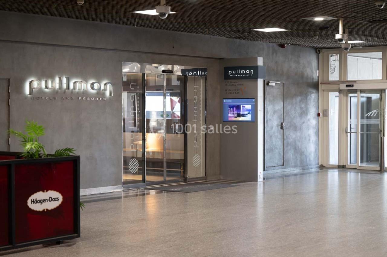 Entrée d'un hôtel Pullman avec portes vitrées, enseigne lumineuse et un écran d'information à droite.