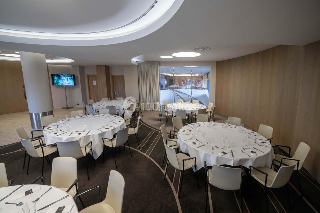 Salle de réception moderne avec tables rondes dressées, chaises blanches et éclairage doux.