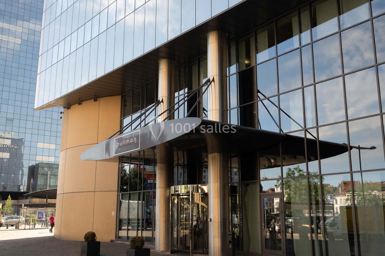 Entrée principale d'un bâtiment moderne avec façade vitrée et auvent noir, entourée de colonnes métalliques.