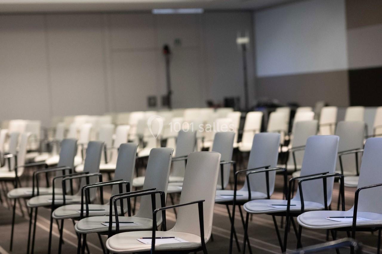 Salle de conférence avec des rangées de chaises blanches et noires alignées, certaines avec des documents posés dessus.