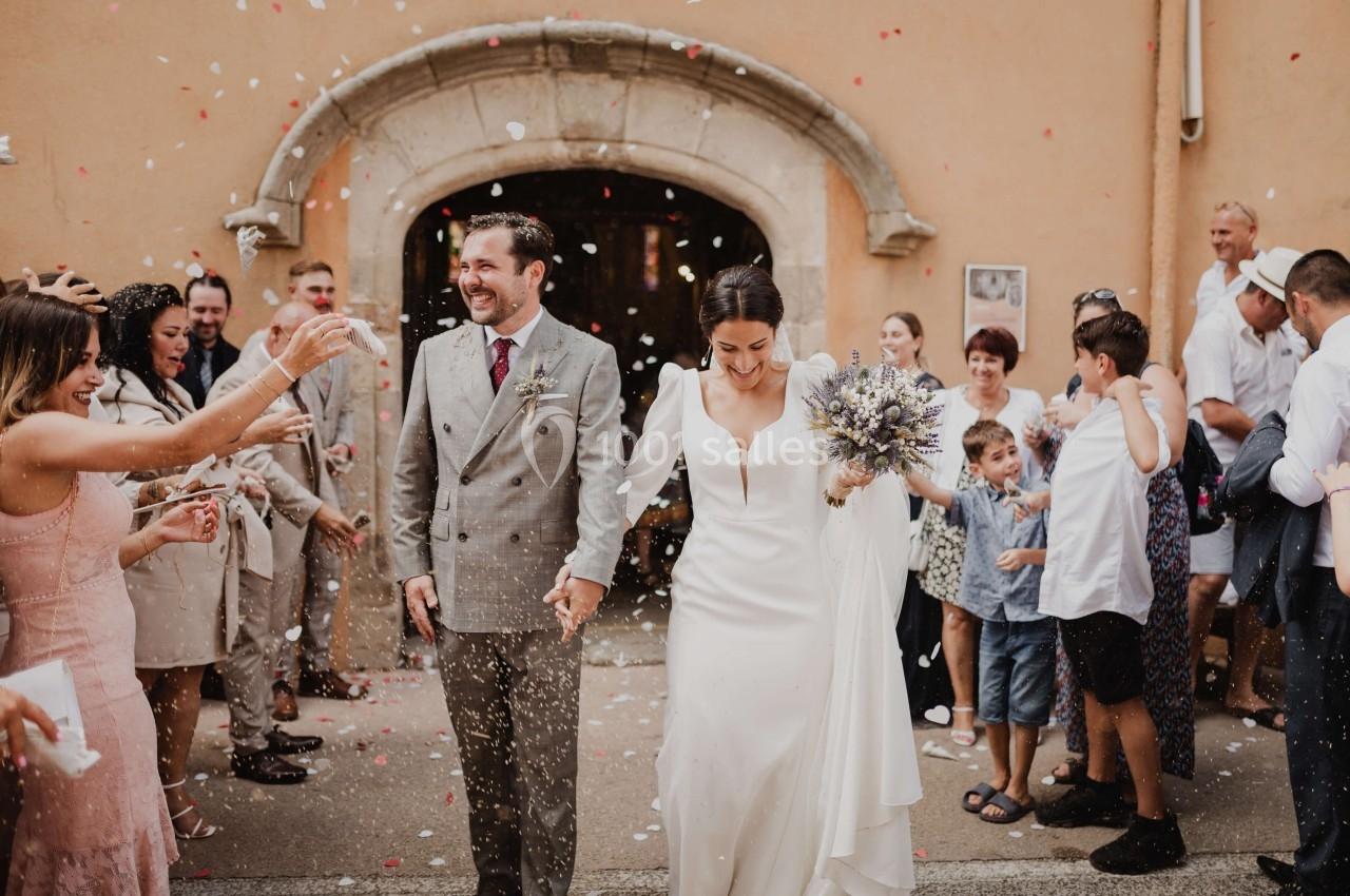 Un couple souriant sort d'un bâtiment sous une pluie de confettis, entouré d'invités célébrant leur mariage.