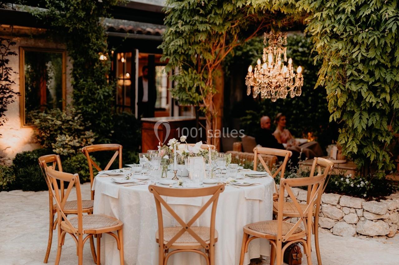 Table ronde dressée avec une nappe blanche, entourée de chaises en bois, dans un jardin éclairé par un lustre suspendu.