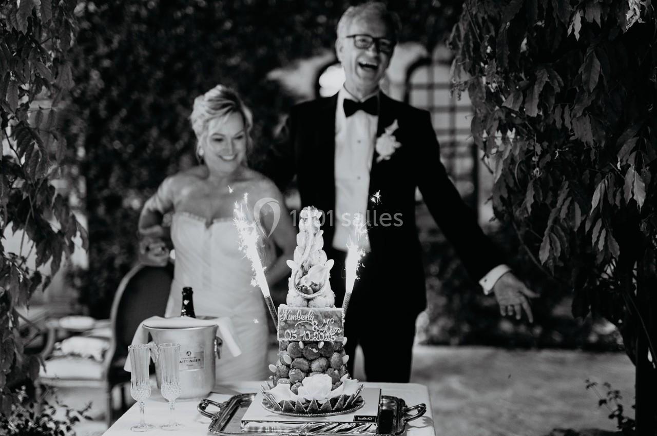 Un couple en tenue de mariage sourit devant un gâteau décoré de feux de Bengale dans un cadre extérieur.