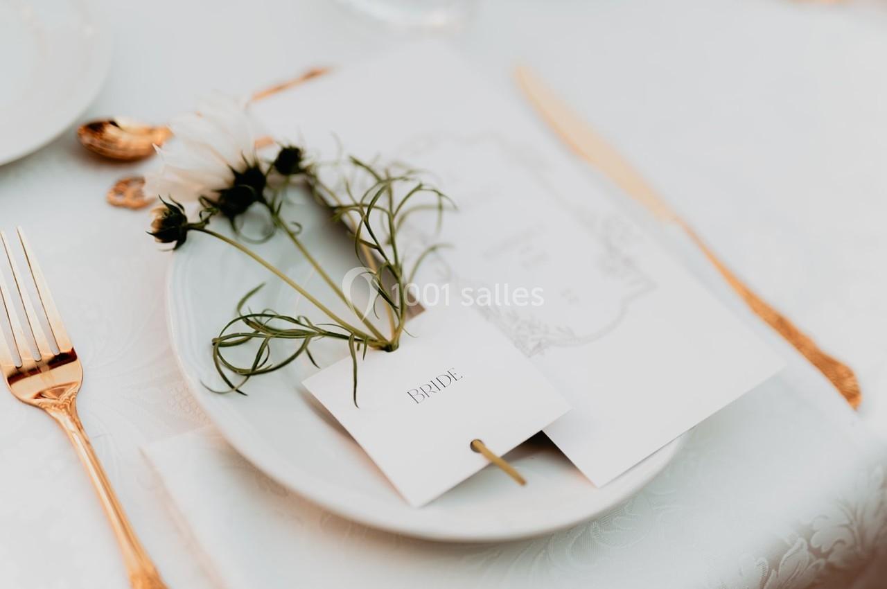 Assiette blanche avec une carte marquée ’Bride’, des fleurs blanches et des couverts dorés sur une nappe claire.