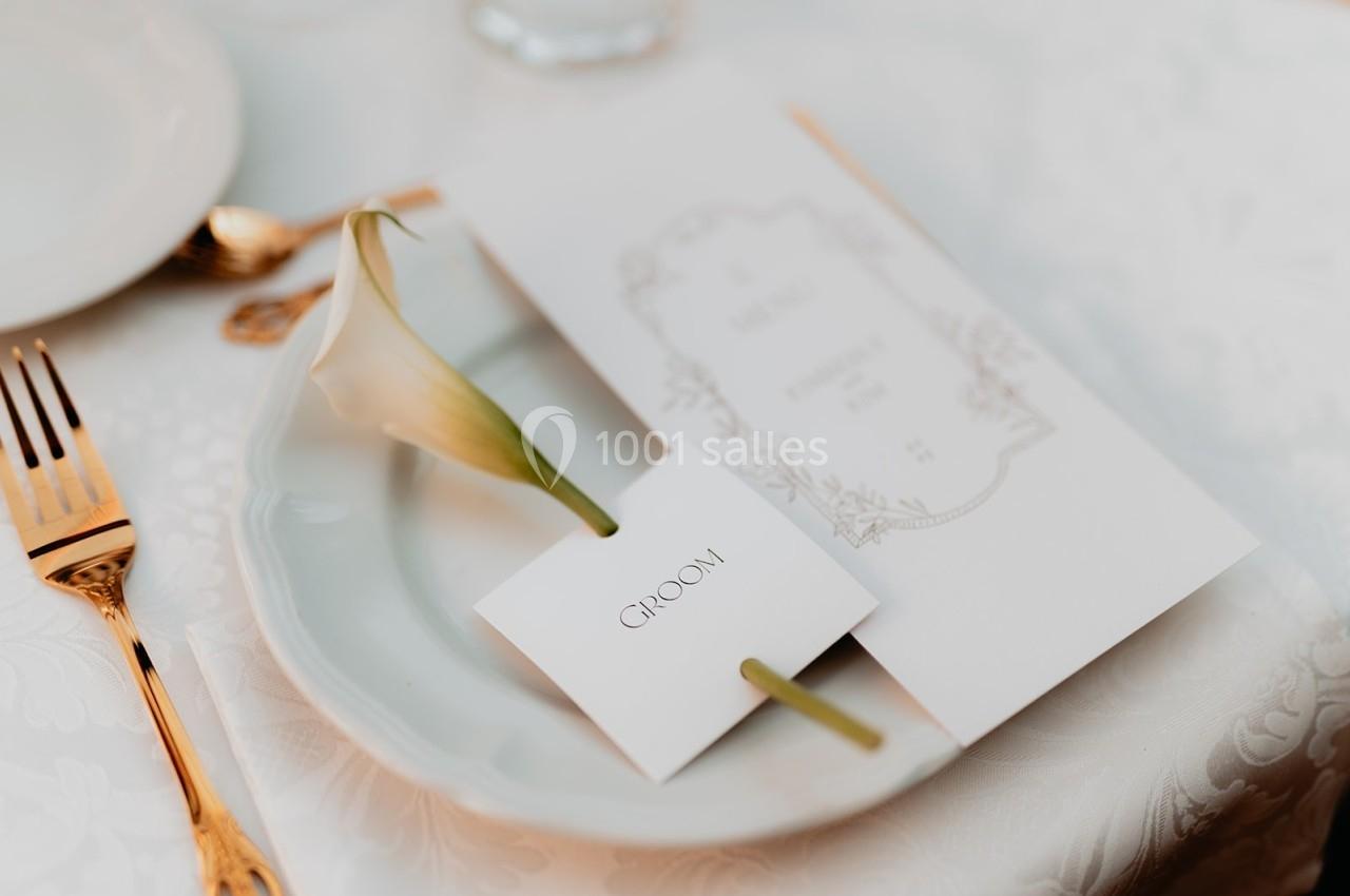 Une assiette blanche avec une fleur de lys, un menu et une carte portant l'inscription ’Groom’ sur une table élégamment…