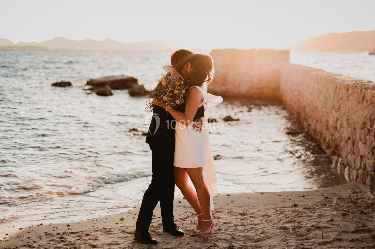 Un couple s'enlace sur une plage au coucher du soleil, près d'un mur en pierre, avec un bouquet de fleurs.
