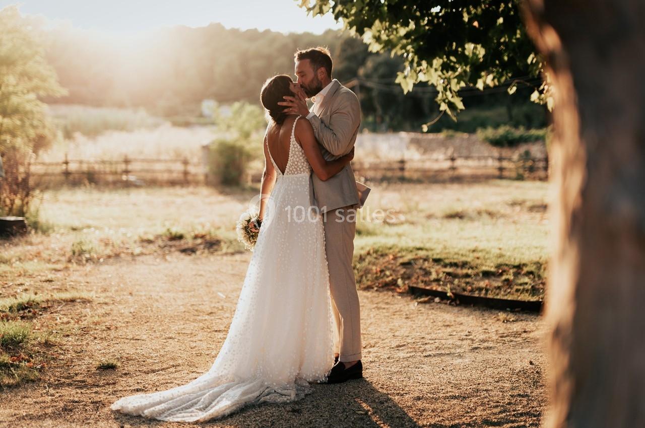 Un couple de mariés s'embrasse tendrement dans un jardin ensoleillé, entouré de nature et d'une clôture en bois.