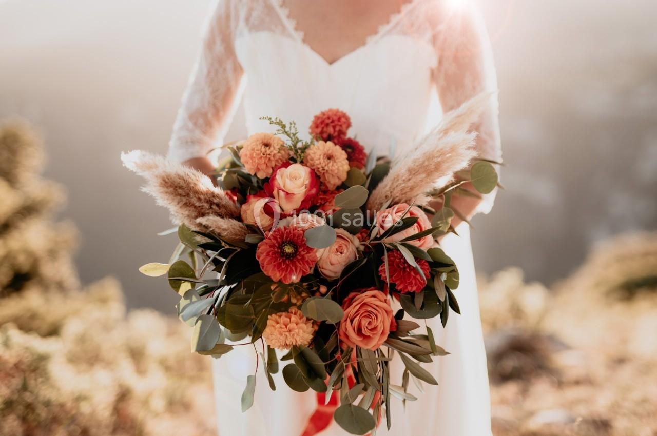 Bouquet de fleurs aux tons chauds avec roses, dahlias et feuillage, tenu par une personne en robe blanche.