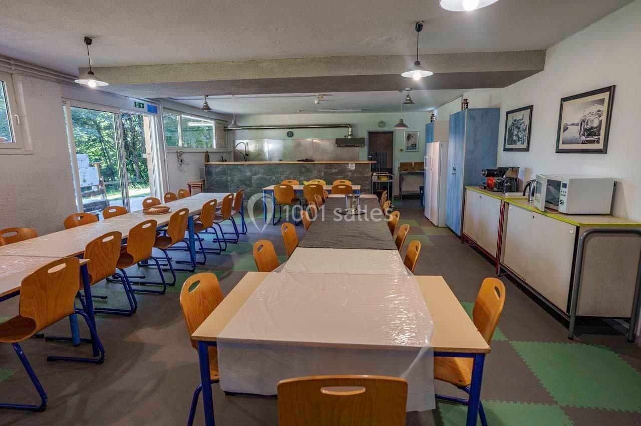 Salle à manger avec tables alignées, chaises en bois, coin cuisine équipé et grandes fenêtres donnant sur l'extérieur.