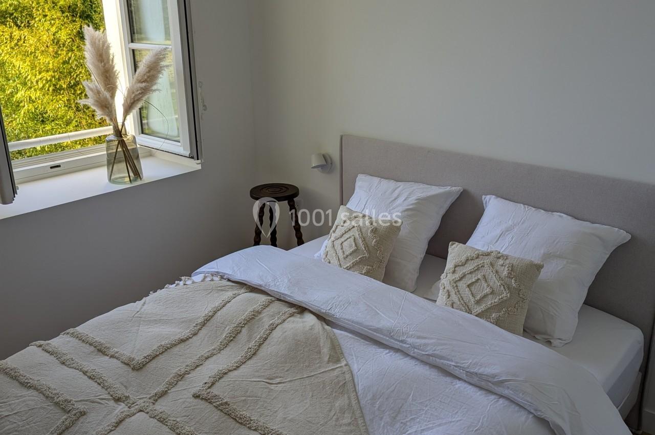 Lit double avec draps blancs et plaid beige, près d'une fenêtre ouverte donnant sur un jardin verdoyant.