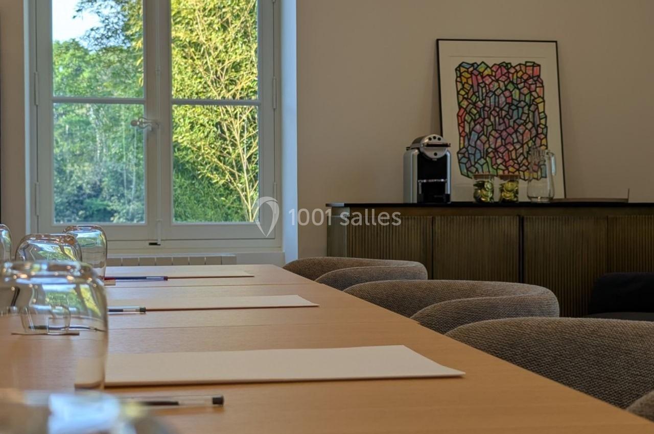 Salle lumineuse avec une table en bois, des feuilles et crayons, vue sur un jardin par une grande fenêtre.