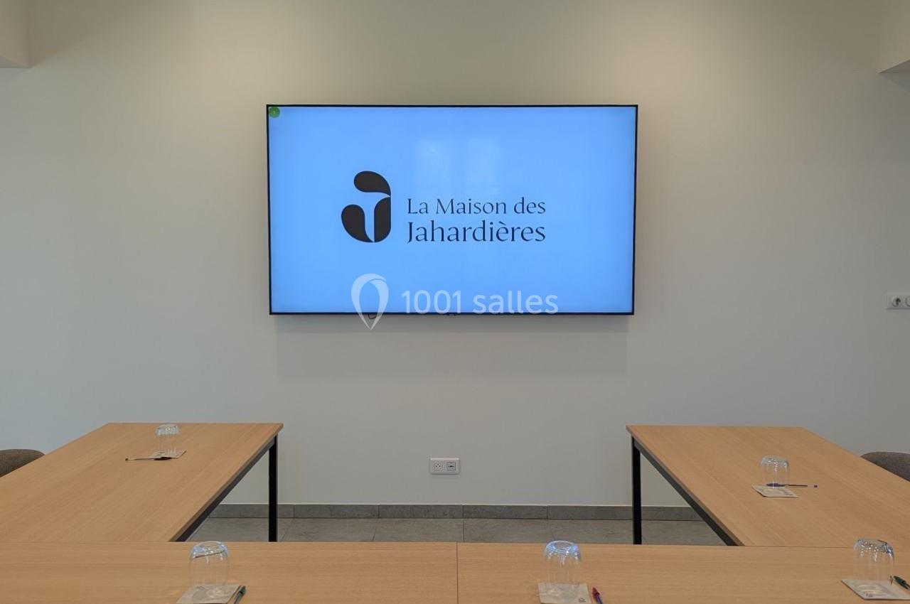 Salle de réunion lumineuse avec tables en U, chaises grises et écran affichant ’La Maison des Jahardières’.