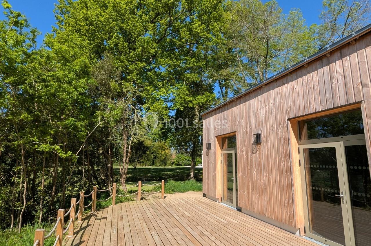 Terrasse en bois bordée de cordes, jouxtant un bâtiment en bois, entourée d'arbres sous un ciel bleu.