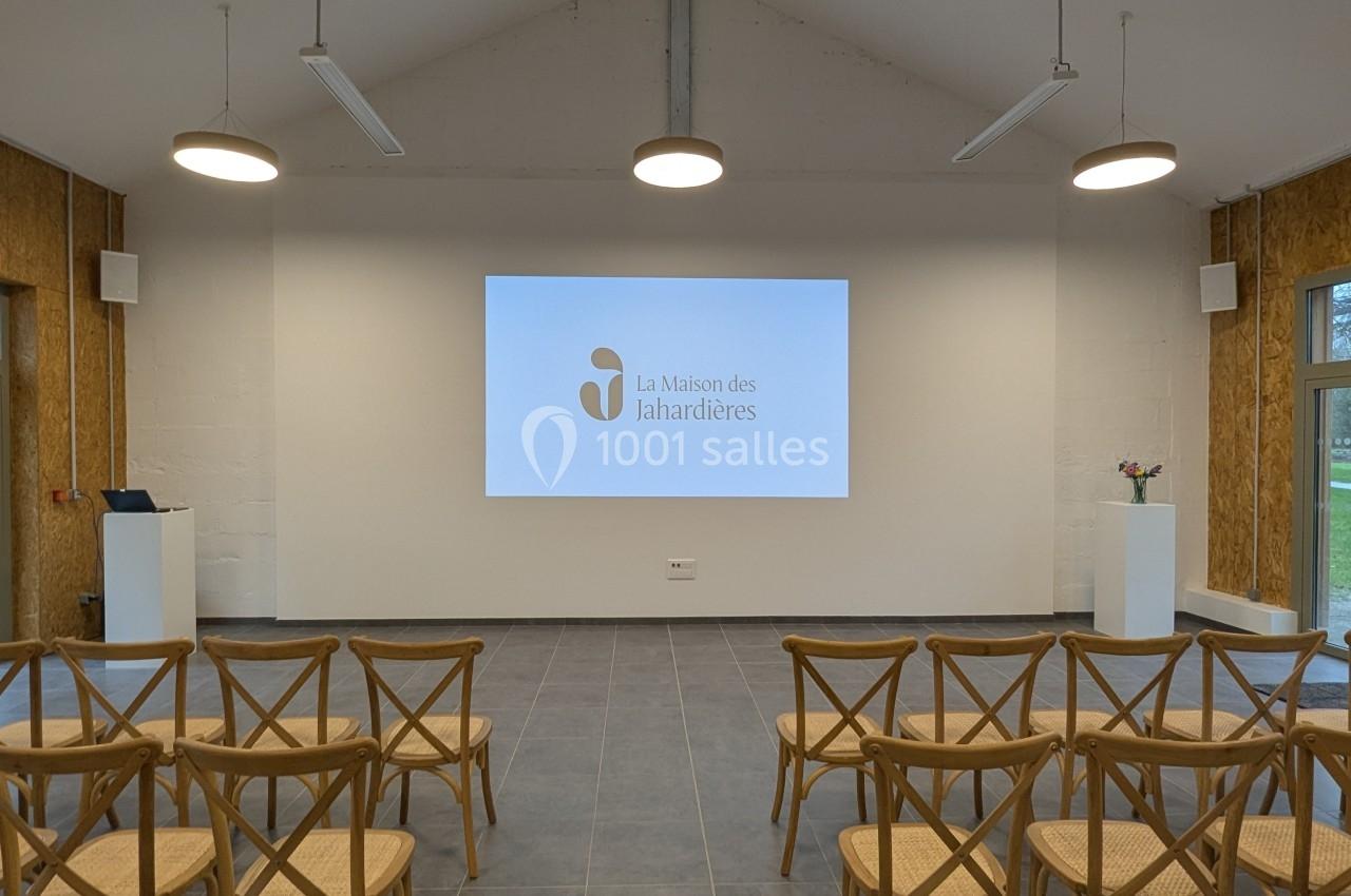 Salle de conférence avec écran de projection allumé, rangées de chaises en bois et éclairage suspendu.