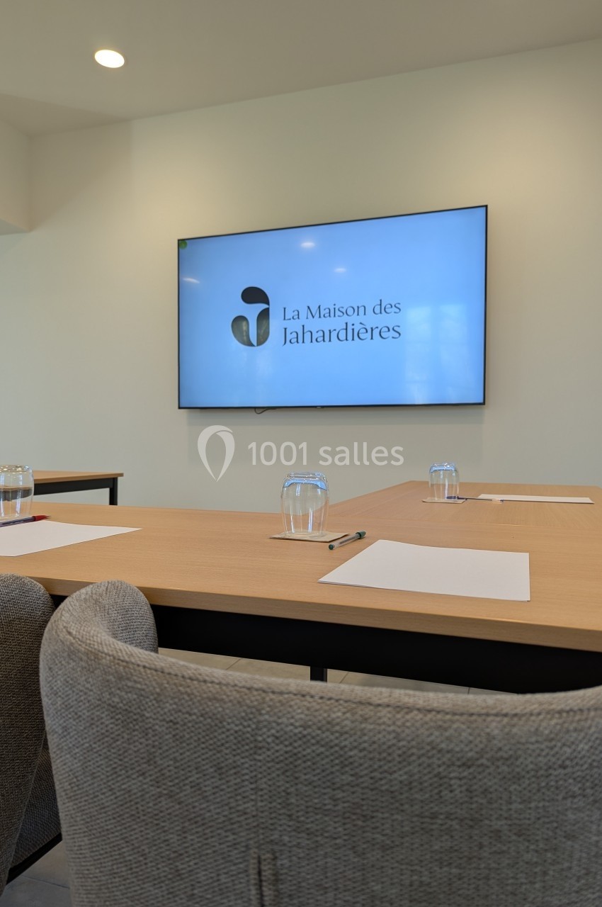 Salle de réunion avec écran affichant le logo ’La Maison des Jahardières’, tables équipées de papiers, stylos et verres.