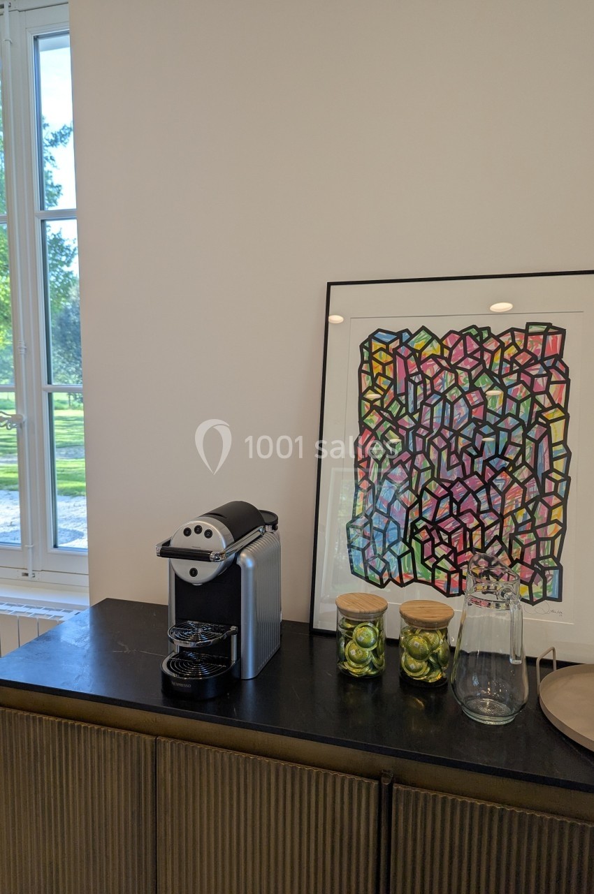 Machine à café sur un buffet avec des bocaux, une carafe et un tableau coloré en arrière-plan près d'une fenêtre.