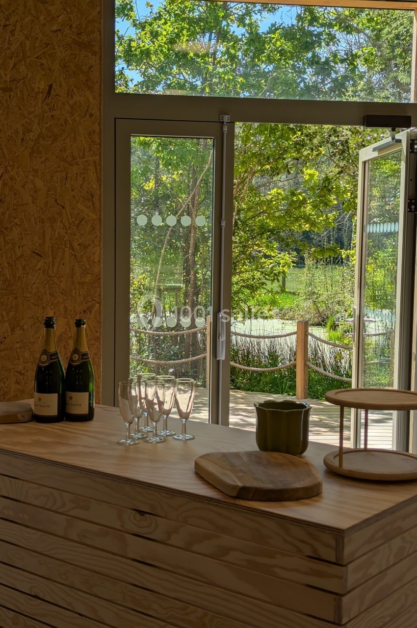 Vue intérieure d'une pièce avec un comptoir en bois, des bouteilles de vin, des verres et une porte ouverte sur un jardin.