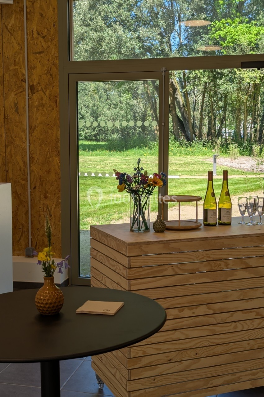 Comptoir en bois avec bouteilles de vin, verres et fleurs, près d'une baie vitrée donnant sur un espace vert.