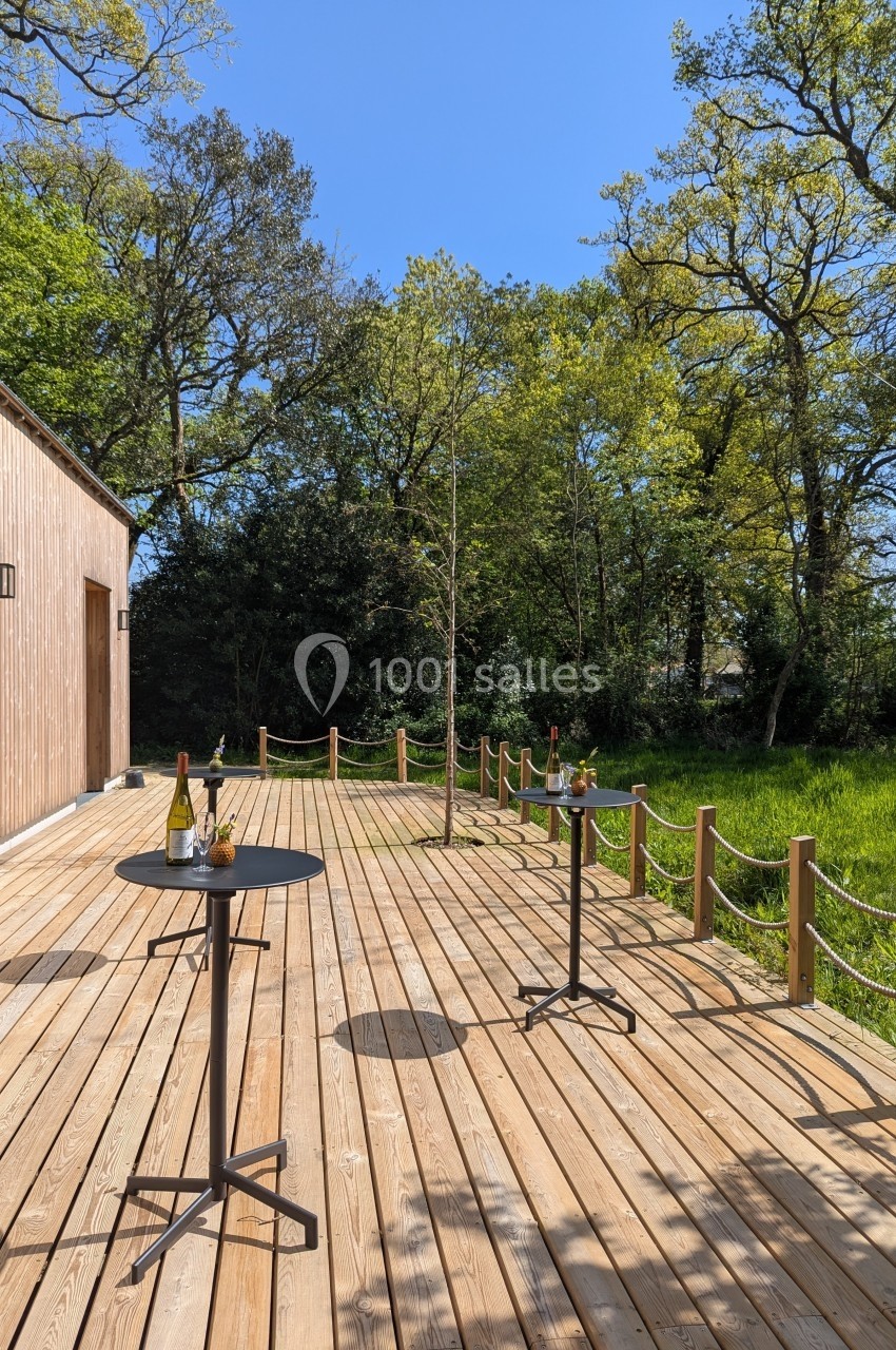 Terrasse en bois avec tables hautes, entourée de verdure et bordée par une corde, sous un ciel bleu clair.