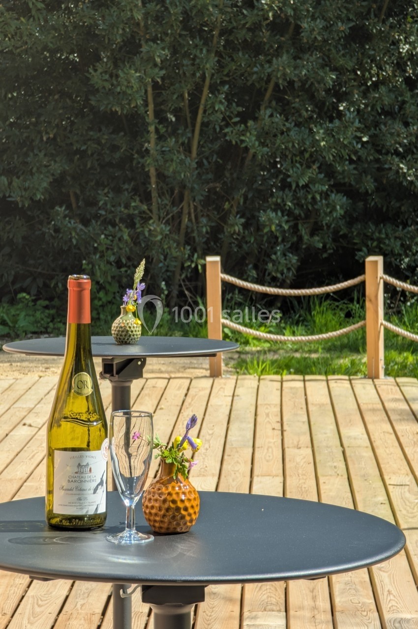 Bouteille de vin, verre et petites décorations florales sur une table ronde en extérieur, avec terrasse en bois et…