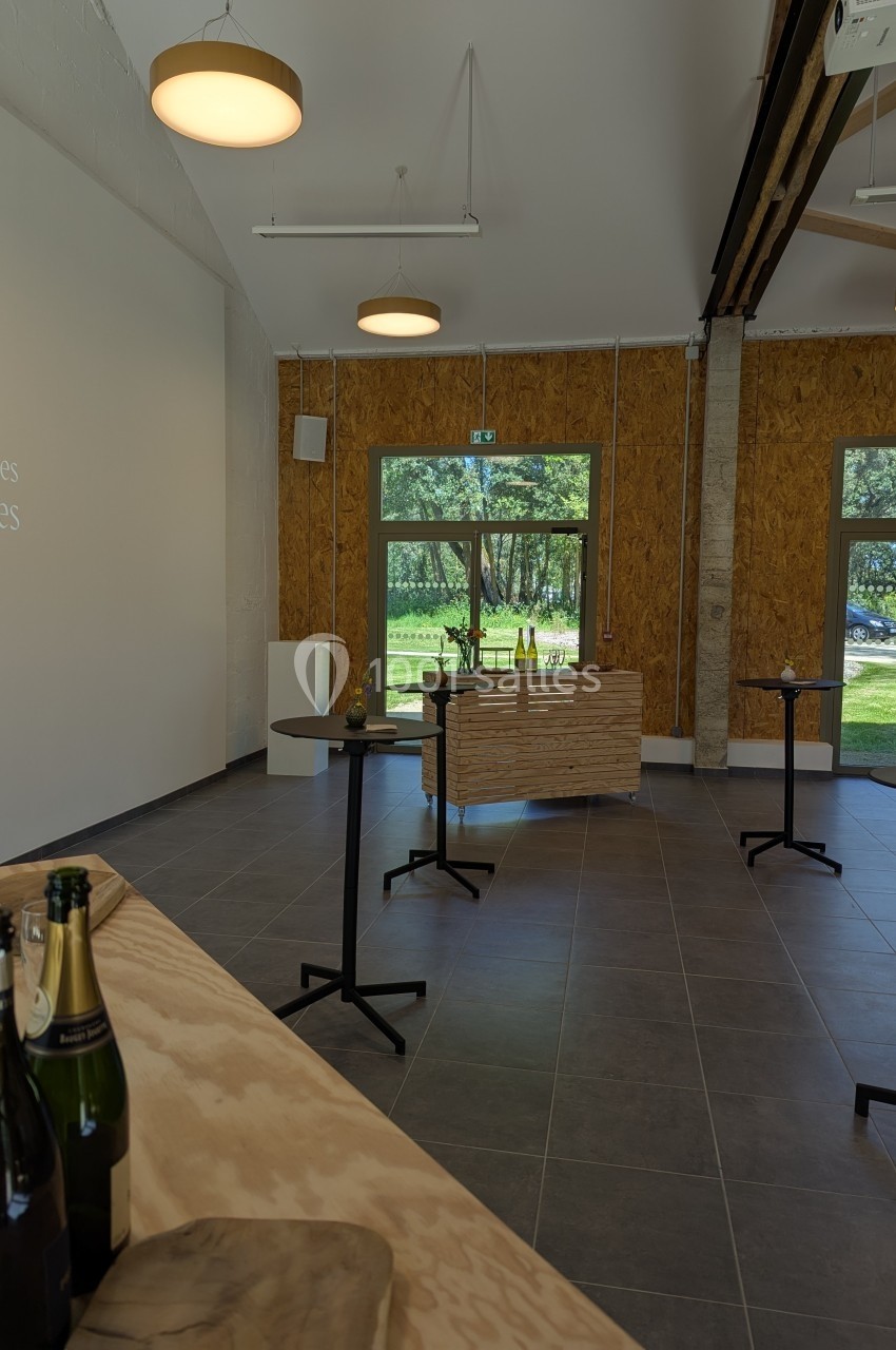 Salle lumineuse avec tables hautes, bouteilles de champagne et vue sur un espace vert à travers de grandes fenêtres.