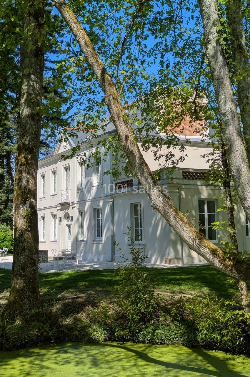 Façade d'une maison blanche entourée d'arbres, avec un petit cours d'eau au premier plan.