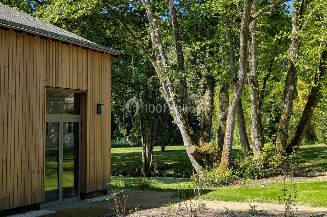Façade en bois d'un bâtiment moderne près d'un jardin arboré avec pelouse et arbres bordant un petit cours d'eau.