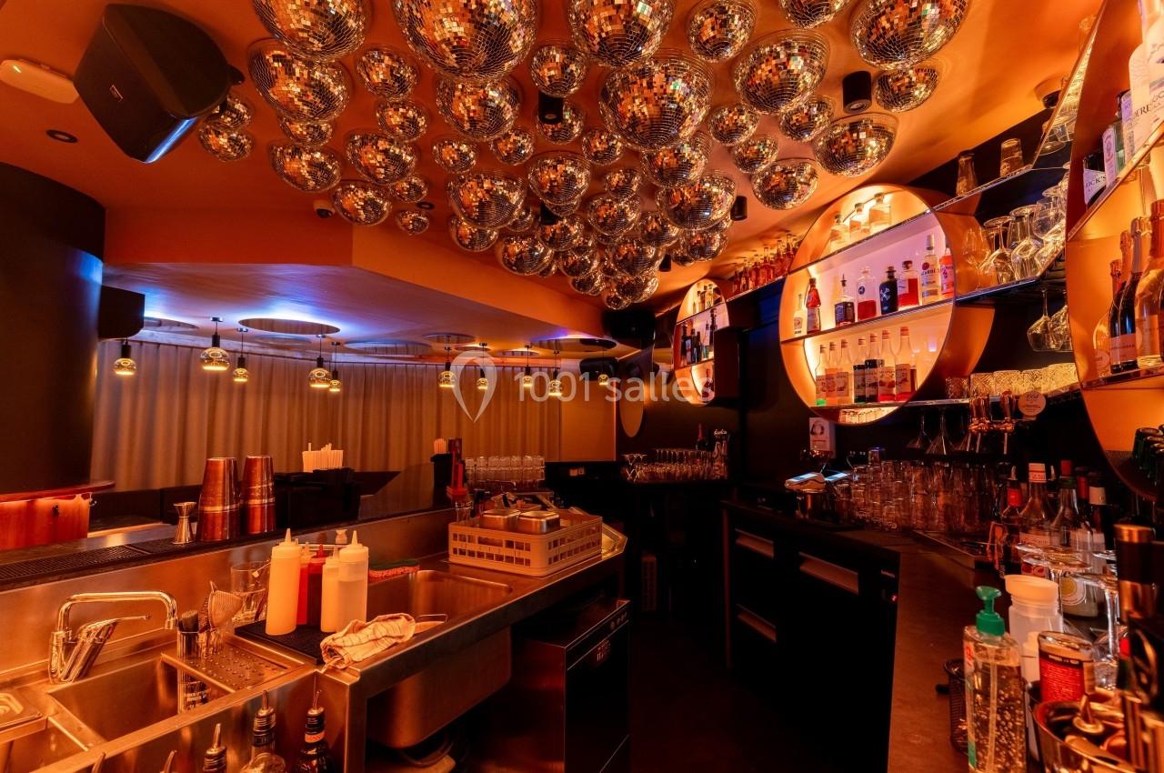 Bar avec comptoir équipé, étagères de bouteilles illuminées et plafond décoré de boules à facettes.