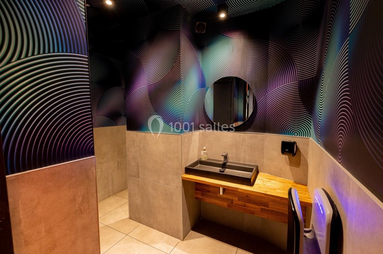 Salle de bain moderne avec lavabo sur un plan en bois, miroir rond et murs décorés de motifs abstraits colorés.