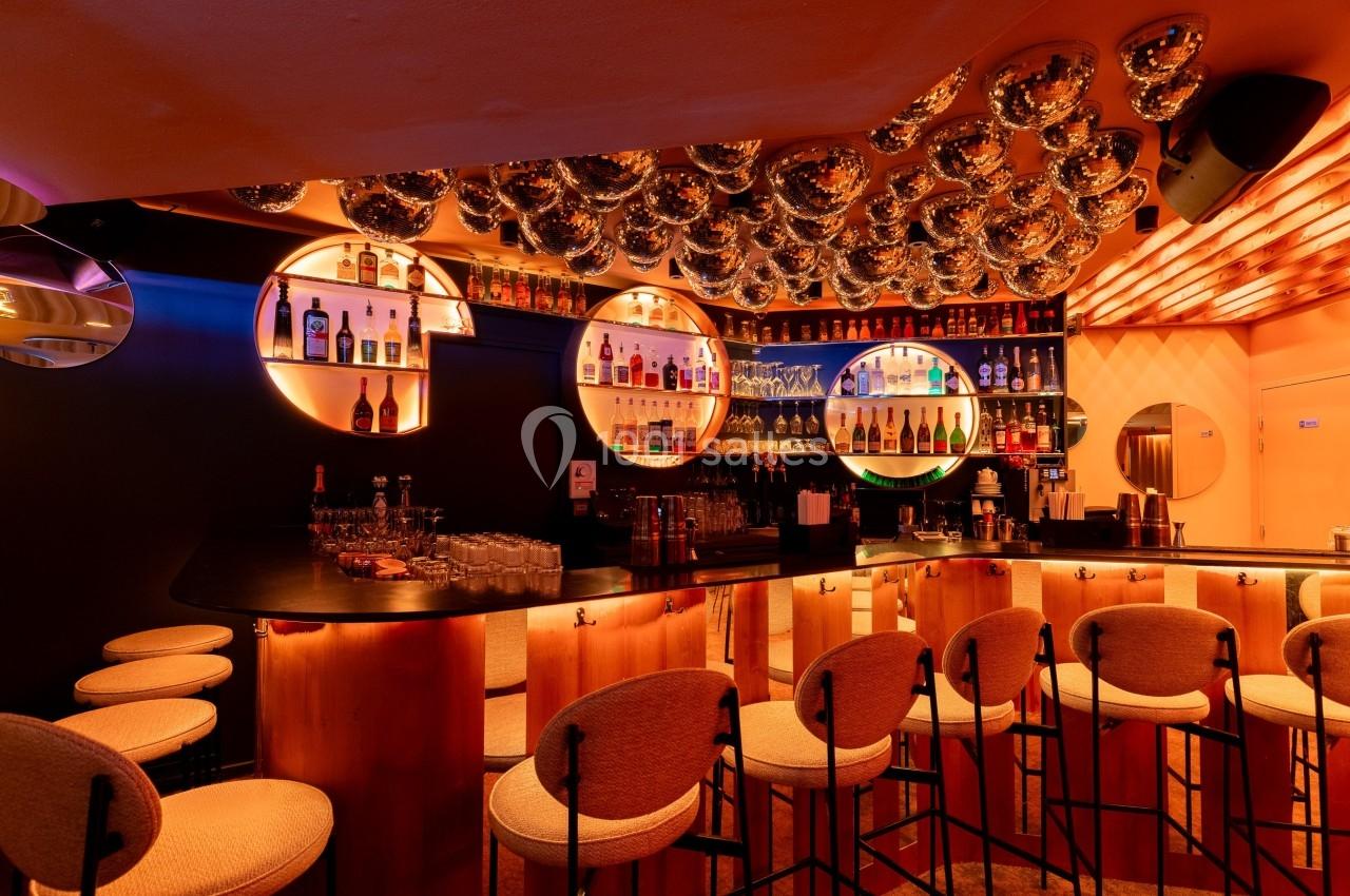 Bar moderne avec comptoir en bois, tabourets rembourrés, étagères éclairées et décorations sphériques au plafond.