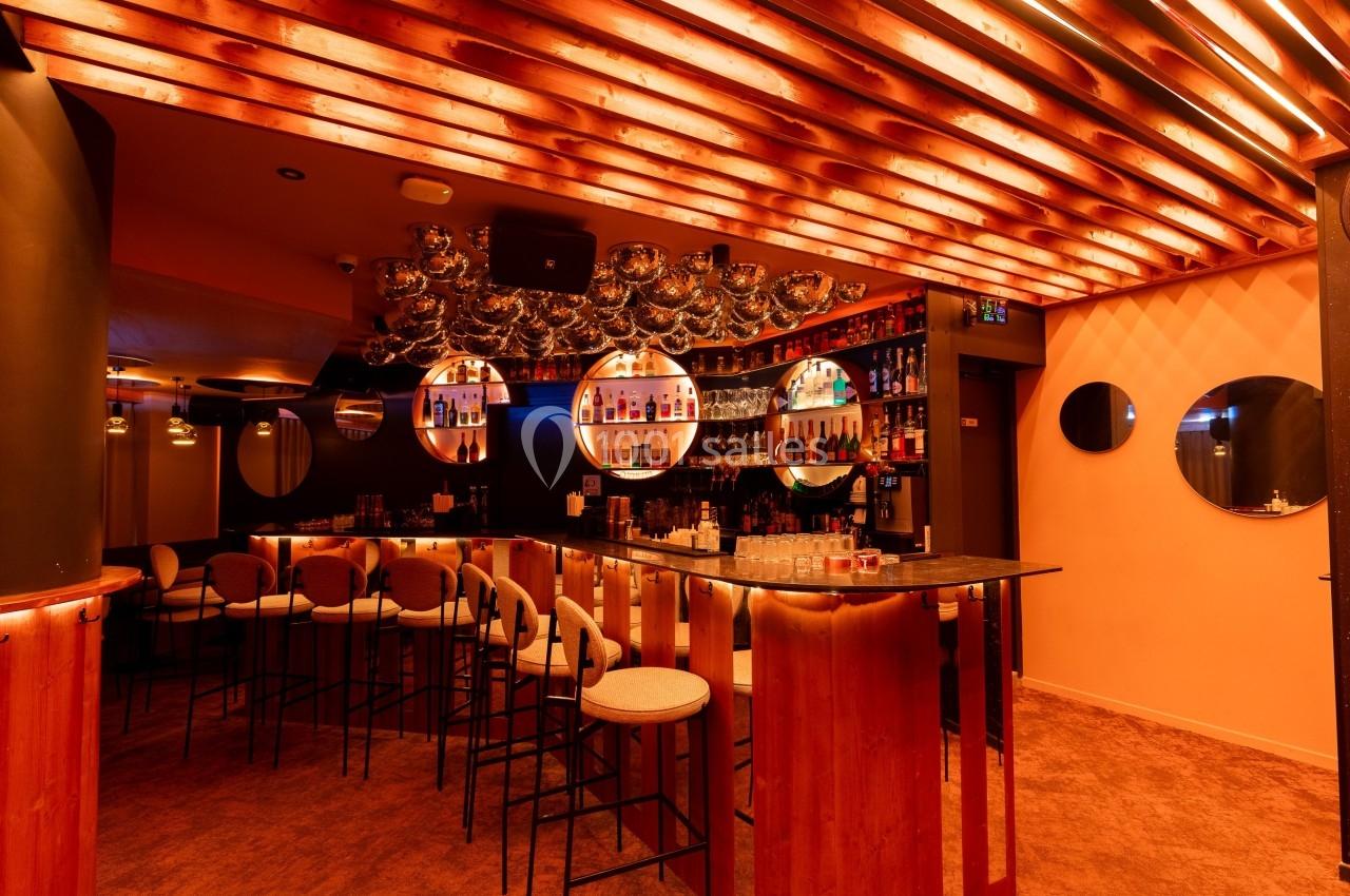 Bar moderne avec comptoir en bois, tabourets hauts, étagères de bouteilles illuminées et éclairage chaleureux.
