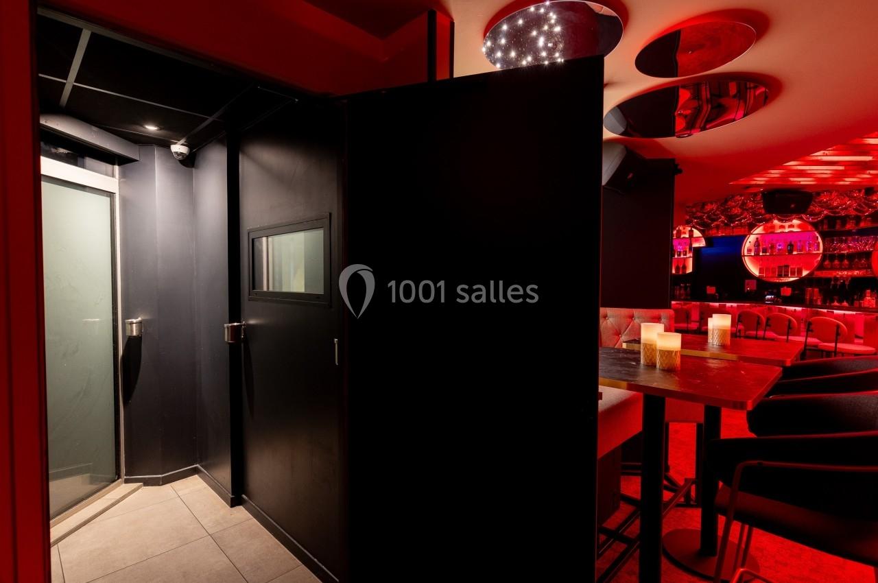 Entrée d'une salle sombre avec porte noire, éclairage rouge et vue partielle sur un bar avec tabourets et tables.