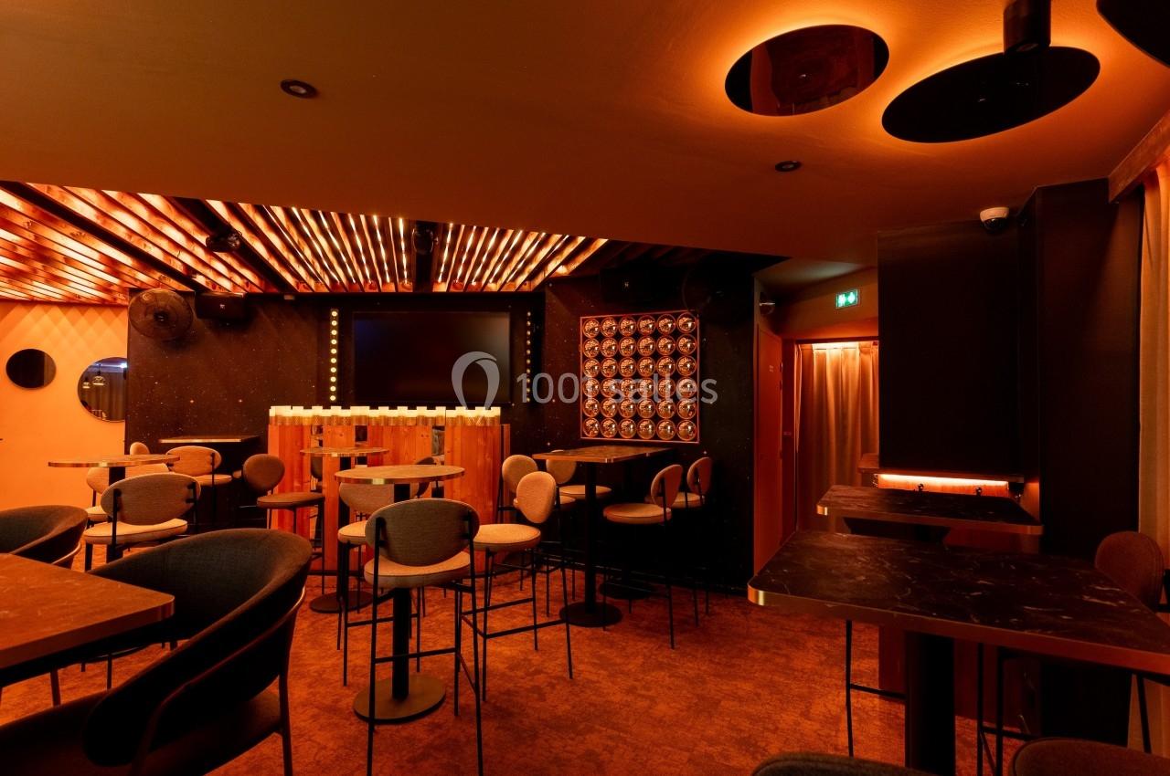 Salle de bar moderne avec éclairage chaleureux, tables hautes, chaises rembourrées et écran mural au fond.