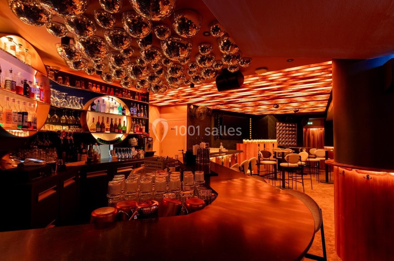 Bar moderne avec comptoir en cuivre, étagères de bouteilles éclairées et plafond décoré de sphères réfléchissantes.