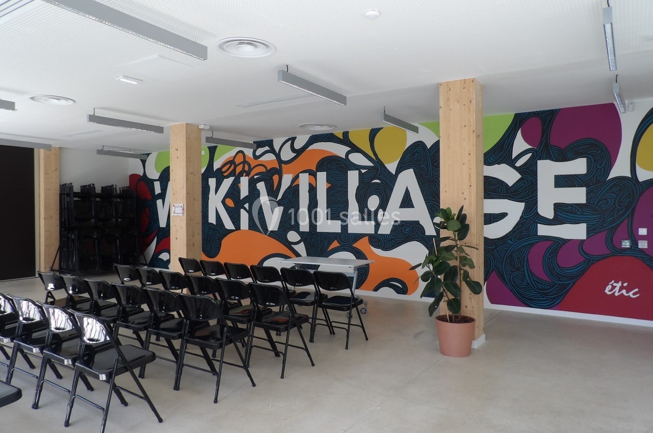Salle avec des chaises noires alignées face à un mur coloré décoré du mot ’Village’ et d'une plante en pot.