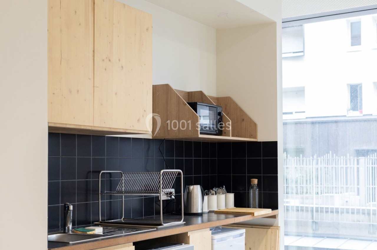 Cuisine moderne avec meubles en bois clair, crédence noire, évier inox, micro-ondes et vue sur l'extérieur.