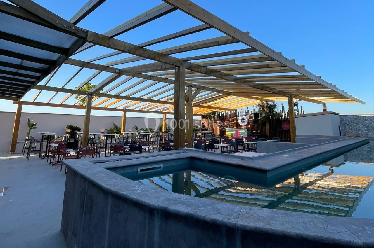 Terrasse extérieure avec tables, chaises et piscine, partiellement couverte par une structure en bois sous un ciel dégagé.