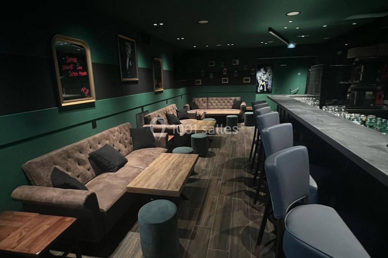 Intérieur d'un bar avec des fauteuils, tables basses, tabourets hauts et un éclairage tamisé dans une ambiance sombre.