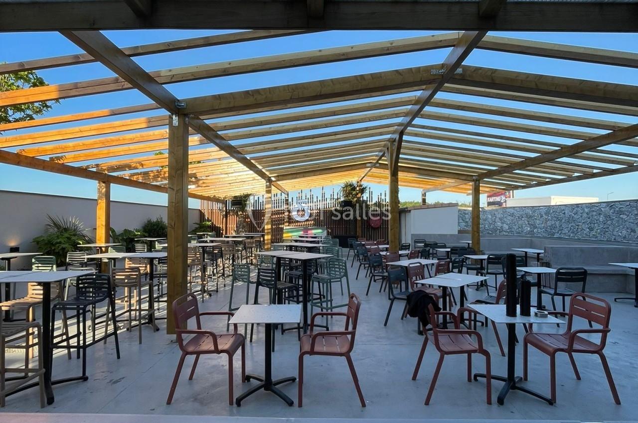 Terrasse extérieure couverte avec tables et chaises espacées, entourée de plantes et éclairée par la lumière naturelle.