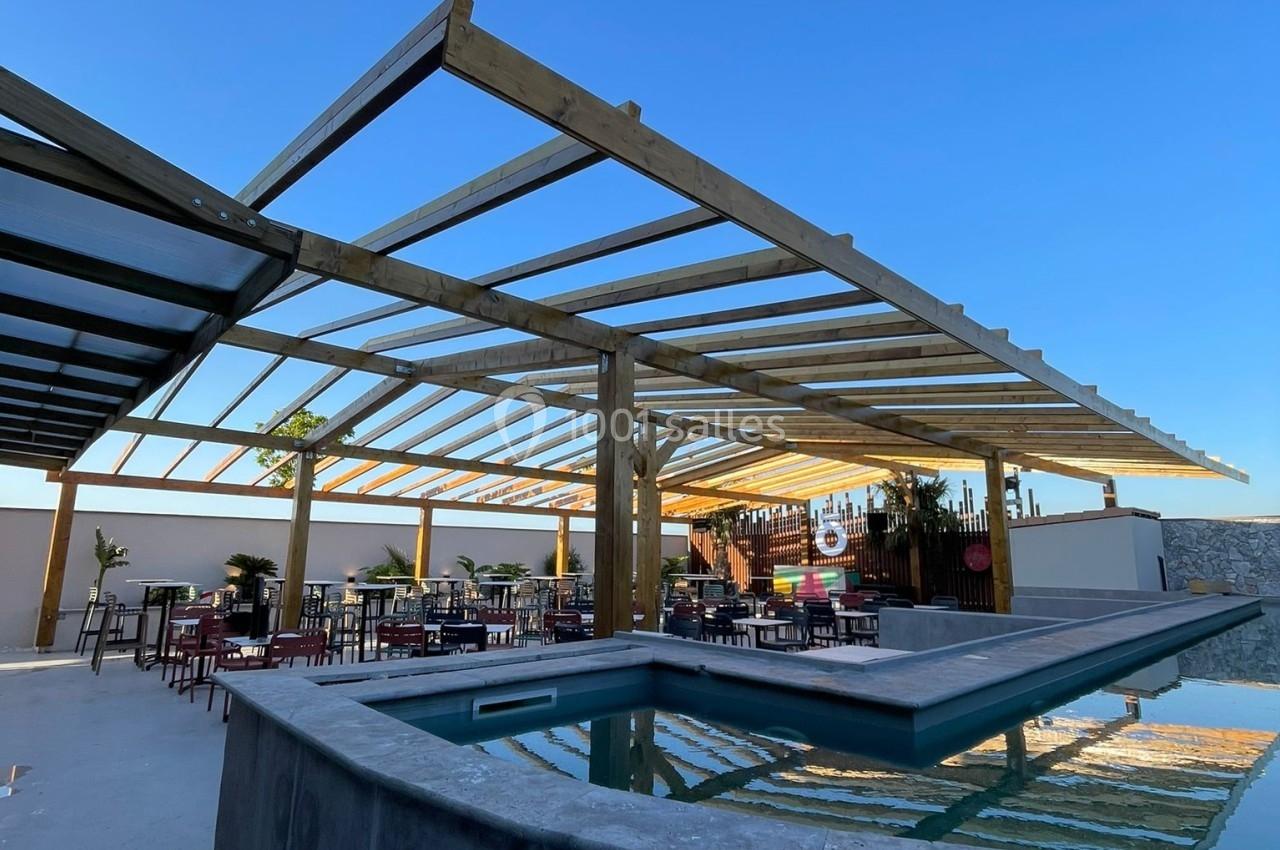 Terrasse extérieure avec piscine, pergola en bois et tables disposées pour un espace de détente sous un ciel dégagé.