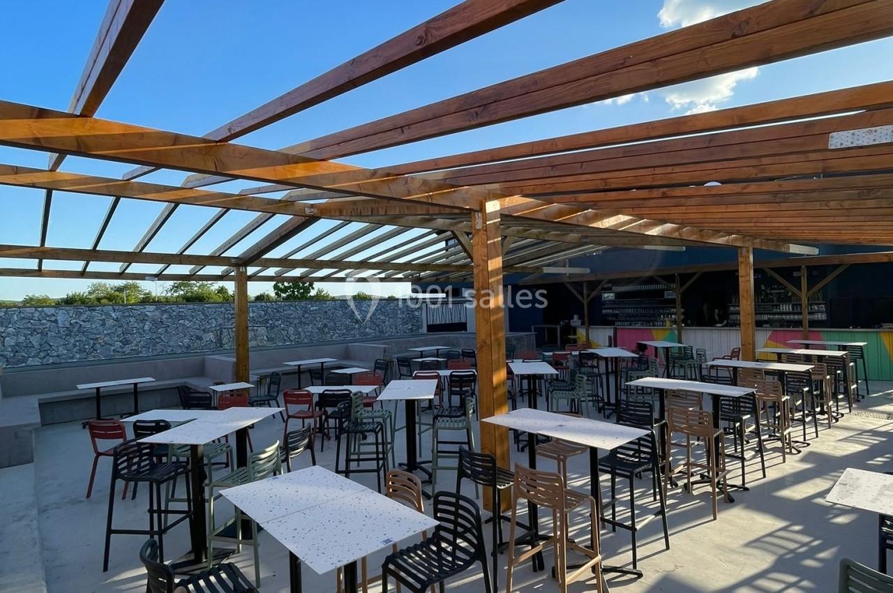 Terrasse extérieure avec tables et chaises sous une structure en bois ouverte, éclairée par un ciel ensoleillé.