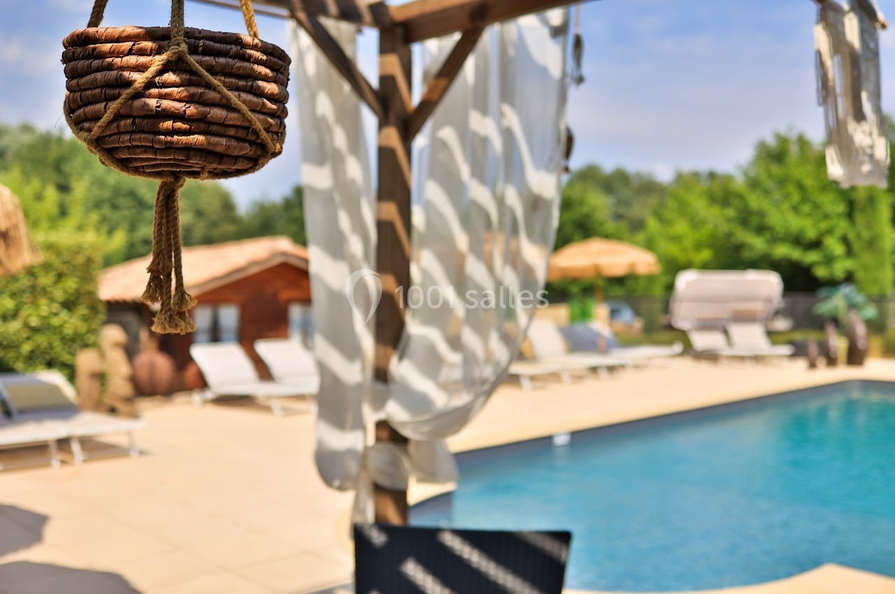 Panier suspendu en osier au premier plan, avec une piscine et des transats floutés en arrière-plan sous un ciel dégagé.