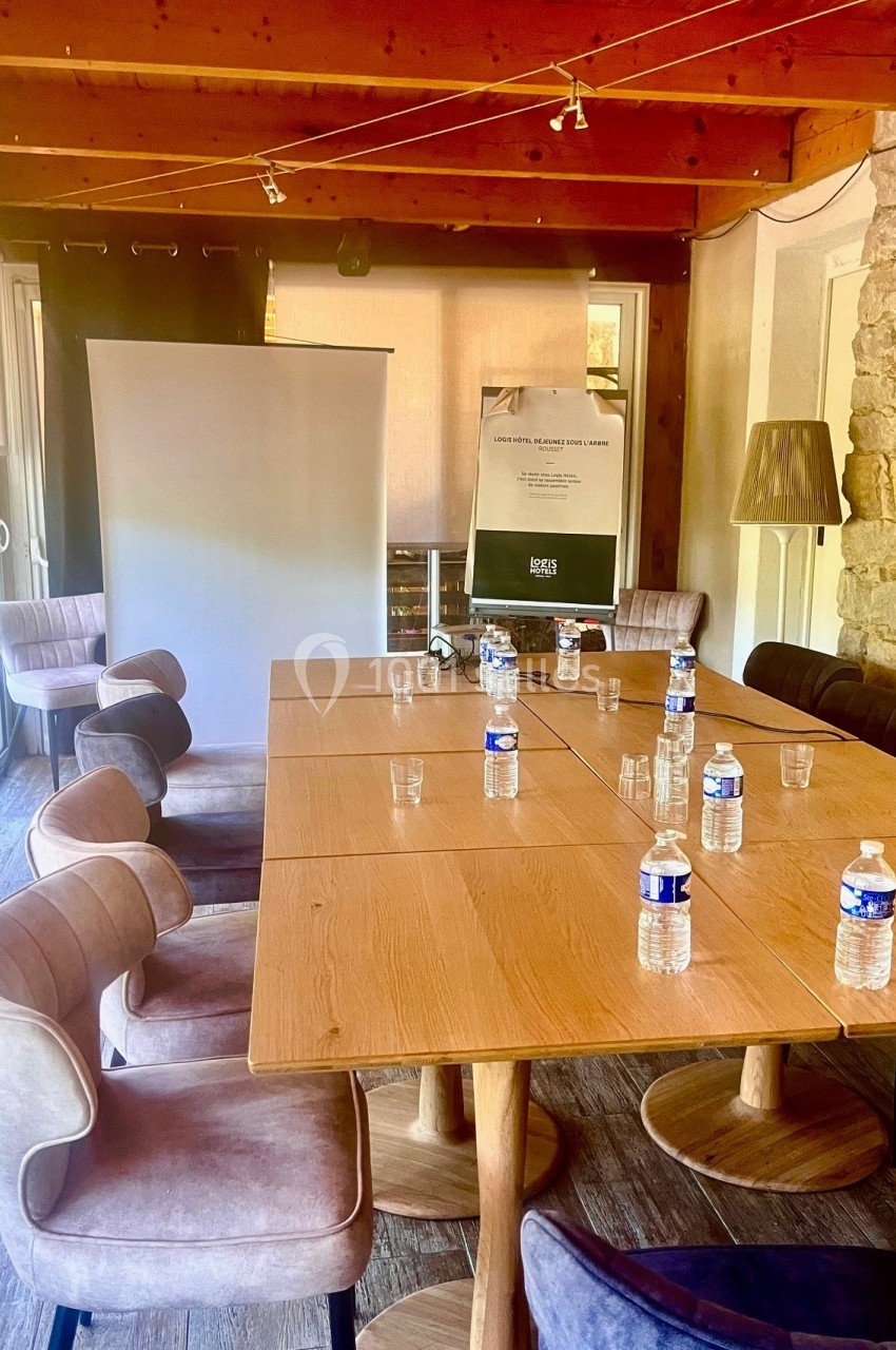 Salle de réunion avec table en bois, chaises rembourrées, bouteilles d'eau, écran de projection et tableau blanc.
