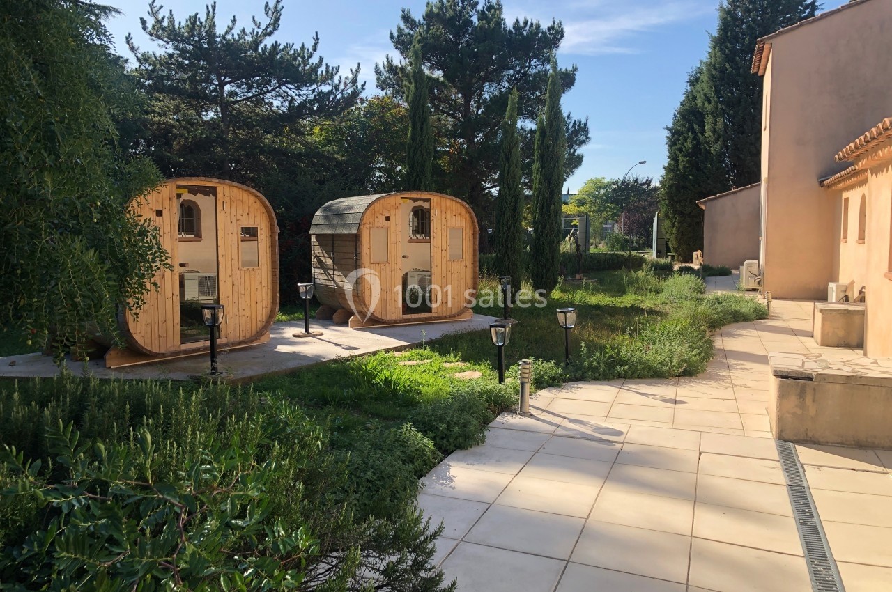 Deux cabanes en bois arrondies dans un jardin verdoyant, près d'une allée pavée et d'un bâtiment beige.