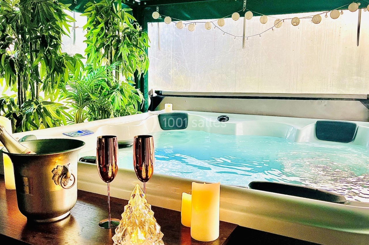 Jacuzzi entouré de plantes, avec bougies, flûtes à champagne et seau à glace sur une table en bois.