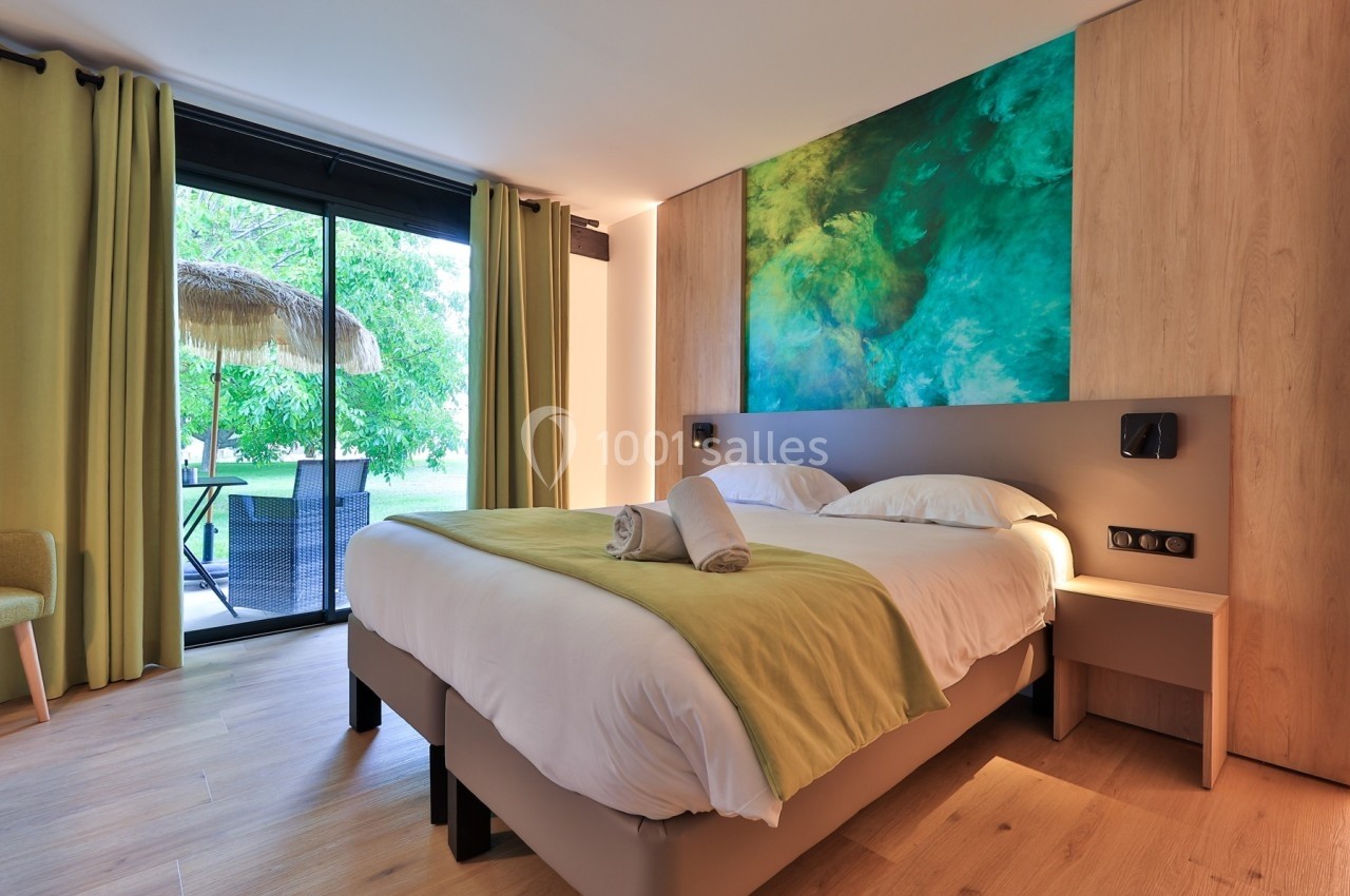 Chambre lumineuse avec lit double, draps blancs, plaid vert, tableau abstrait et accès à une terrasse avec vue sur jardin.